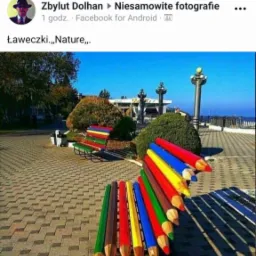 Kolorowa ławka w kształcie kredek na tle parku miejskiego z widokiem na morze w słoneczny dzień.