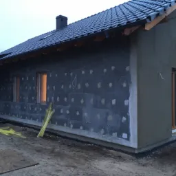 Budowa domu jednorodzinnego w Poznaniu na etapie ocieplania ścian zewnętrznych, widoczne bloczki oraz częściowo nałożony tynk. Na pierwszym planie leżą żółte listwy i deska.