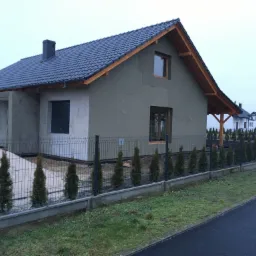 Surowy, otynkowany dom jednorodzinny w trakcie budowy, z ciemną dachówką, widoczny od frontu, z ogrodzeniem z siatki i młodymi tujami wzdłuż niego, na tle pochmurnego nieba.