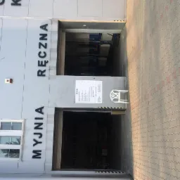 Fasada budynku z napisem 'MYJNIA RĘCZNA', widoczne otwarte wrota wjazdowe, obok cenik i plastikowe krzesło.