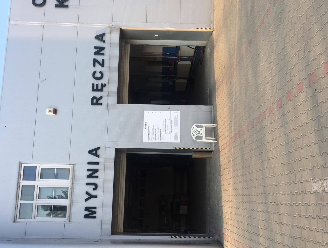 Fasada budynku z napisem 'MYJNIA RĘCZNA', widoczne otwarte wrota wjazdowe, obok cenik i plastikowe krzesło.