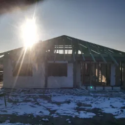 Surowy stan domu murowanego z widoczną konstrukcją dachu w zimowej scenerii, silne słońce w kadrze.