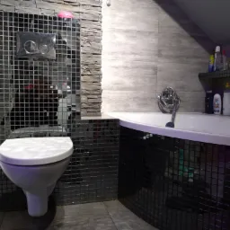 Nowoczesna łazienka z toaletą i wanną obłożona czarną mozaiką, ściana z szarej płytki imitującej kamień, półka z kosmetykami w tle.