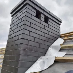 Nowy, ciemnoszary komin z cegły na dachu w trakcie budowy, widoczne drewniane elementy konstrukcji dachu i membrana dachowa.