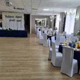 Sala bankietowa z dekoracją na przyjęcie: ciemnoniebieskie obrusy, białe krzesła z pokrowcami, kwiatowe kompozycje na stołach i dekoracja ściany z napisem 'Najlepsze dopiero przed nami'.