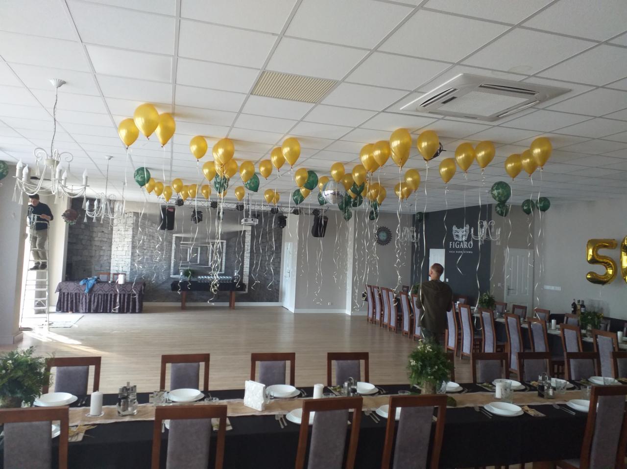 Przygotowania do uroczystości: złote i zielone balony pod sufitem, dekoracja sali z ustawionymi stołami, pracownik na drabinie montujący dekoracje, napis 'Figaro' na ścianie.
