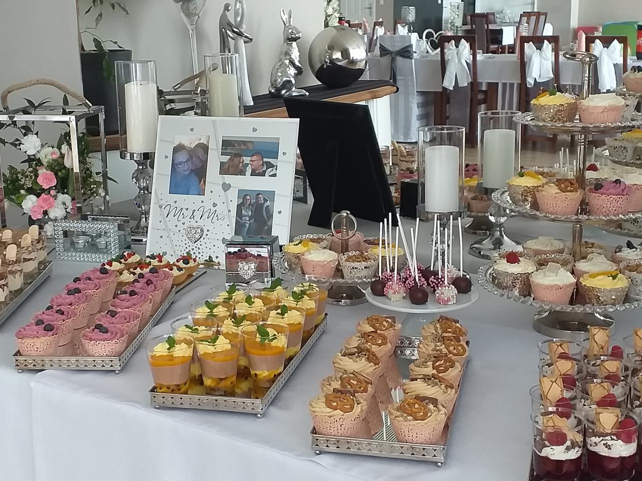 Stół z deserami na przyjęciu: babeczki z preclami, musy w szklankach, cake pops, patery z ciastkami, świece w szklanych świecznikach, ramka ze zdjęciami pary młodej, srebrne figurki dekoracyjne.