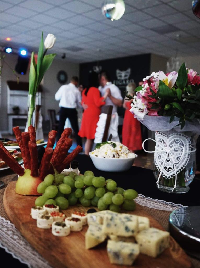 Stół z przekąskami: winogrona, ser pleśniowy, koreczki, kabanosy wbite w jabłko, sałatka w miseczce, bukiet kwiatów w wazonie z koronkowym sercem, w tle tańcząca para na sali balowej.