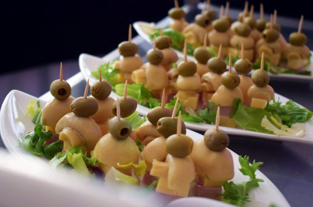 Elegancko podane koreczki na białych talerzach: oliwki, pieczarki, ser i szynka na zielonej sałacie.