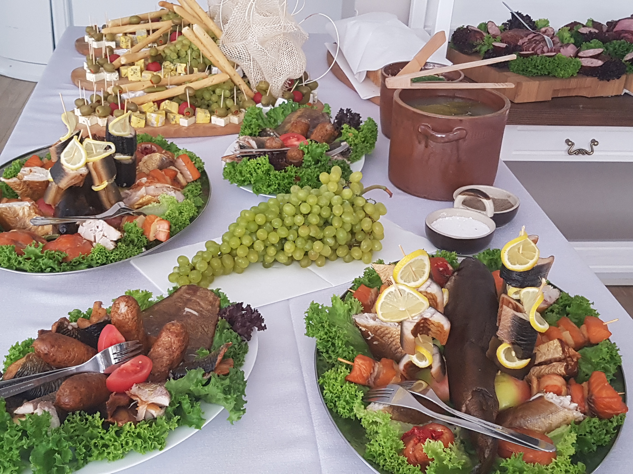 Stół z różnorodnymi potrawami: wędzone ryby z cytryną, koreczki z serem i oliwkami, sałatki, waza z zupą, wszystko udekorowane zieloną sałatą, z widocznymi sztućcami.