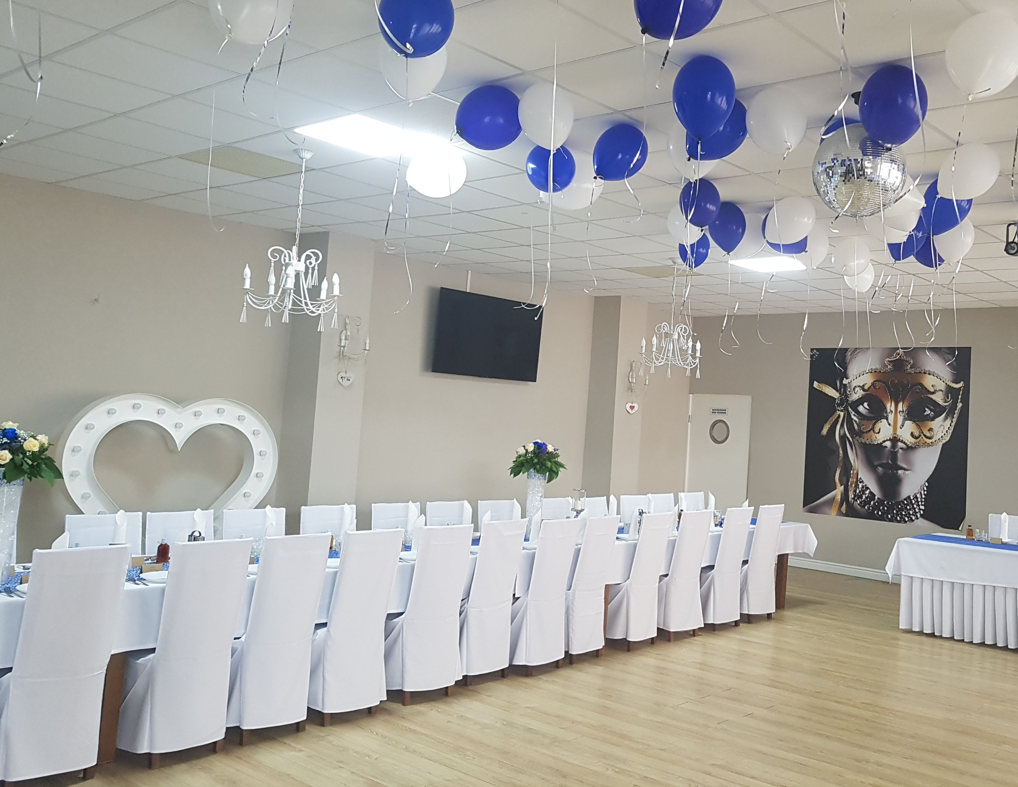 Sala udekorowana na imprezę: białe krzesła z pokrowcami, długi stół z niebieskim bieżnikiem, balony białe i niebieskie pod sufitem, lustrzana kula, dekoracja w kształcie serca z żarówkami...