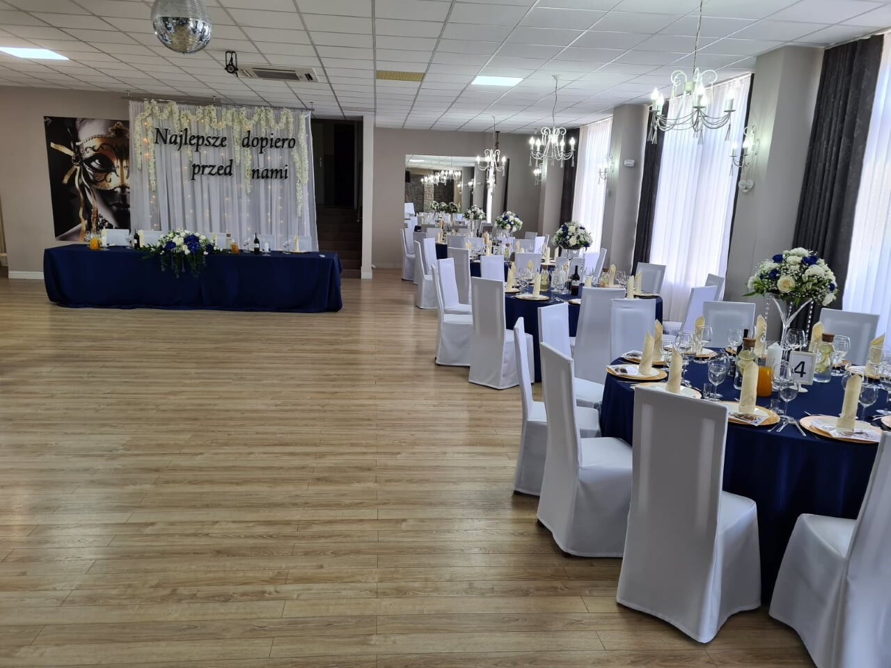 Sala bankietowa z dekoracją na przyjęcie: ciemnoniebieskie obrusy, białe krzesła z pokrowcami, kwiatowe kompozycje na stołach i dekoracja ściany z napisem 'Najlepsze dopiero przed nami'.