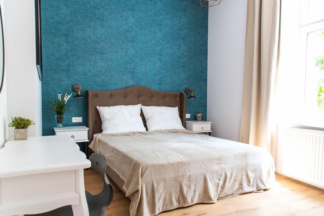 Jasna sypialnia z turkusową ścianą, brązowym łóżkiem z białymi poduszkami, białym biurkiem i drewnianą podłogą, oświetlona lampkami ściennymi.