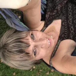 Uśmiechnięta kobieta z blond włosami leży na trawie w hamaku, patrząc w górę. Widoczne liście na trawie i wzorzysta sukienka.