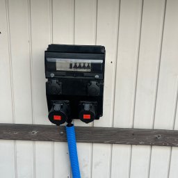 SPIDER INSTAL Piotr Zawadzki - Czarna skrzynka elektryczna z gniazdami siłowymi i niebieską rurą karbowaną, zamontowana na białej, drewnianej ścianie z poziomą deską. Widoczne zabezpieczenia w skrzynce.