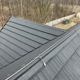 SPIDER INSTAL Piotr Zawadzki - Czarny dach z blachy na rąbek stojący z zamontowaną instalacją odgromową, widok z góry na tle drzew i ogrodzenia. Detal mocowania przewodów odprowadzających.