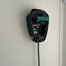 SPIDER INSTAL Piotr Zawadzki - Rozmontowana, czarna stacja ładowania EV na białej ścianie, z widocznymi wewnętrznymi obwodami i okablowaniem. Biały czujnik alarmowy w tle, obok framugi drzwi.