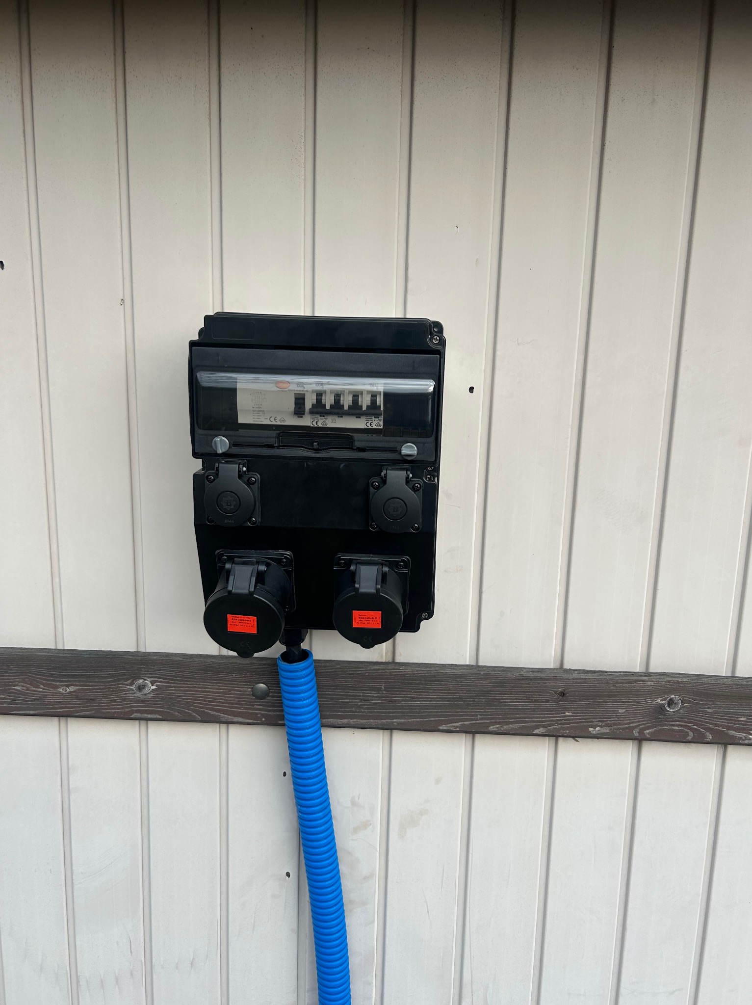 Czarna skrzynka elektryczna z gniazdami siłowymi i niebieską rurą karbowaną, zamontowana na białej, drewnianej ścianie z poziomą deską. Widoczne zabezpieczenia w skrzynce.