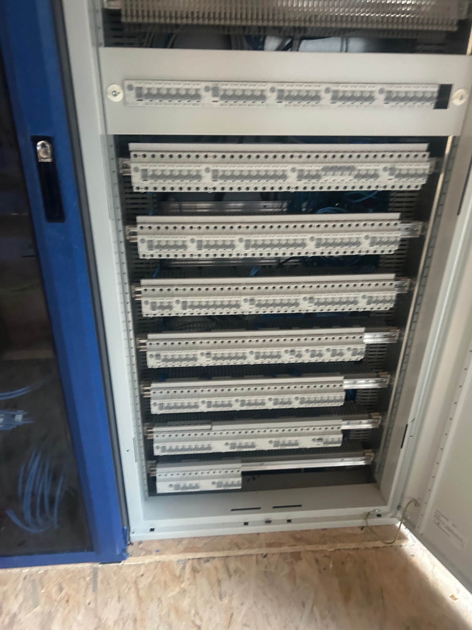 Otwarta szafa sterownicza z licznymi listwami zaciskowymi i kablami, widoczna drewniana podłoga i fragment niebieskiej obudowy. Ujęcie z bliska.