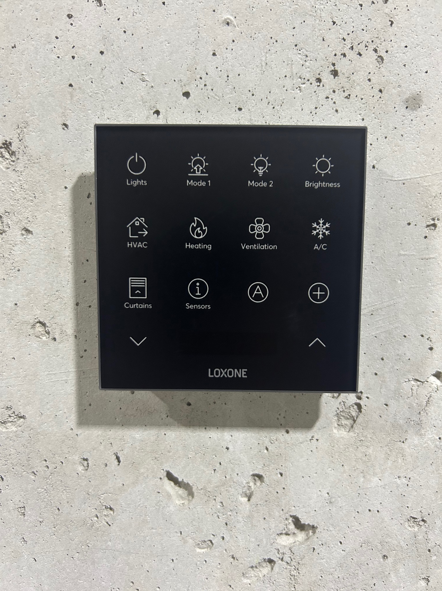 Czarny panel dotykowy Loxone na szarej, surowej ścianie betonowej. Sterowanie oświetleniem, klimatyzacją, ogrzewaniem, wentylacją, roletami i innymi funkcjami domu.