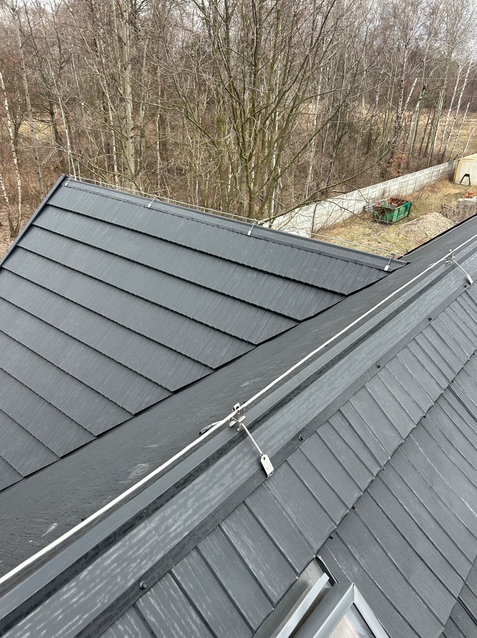 Czarny dach z blachy na rąbek stojący z zamontowaną instalacją odgromową, widok z góry na tle drzew i ogrodzenia. Detal mocowania przewodów odprowadzających.