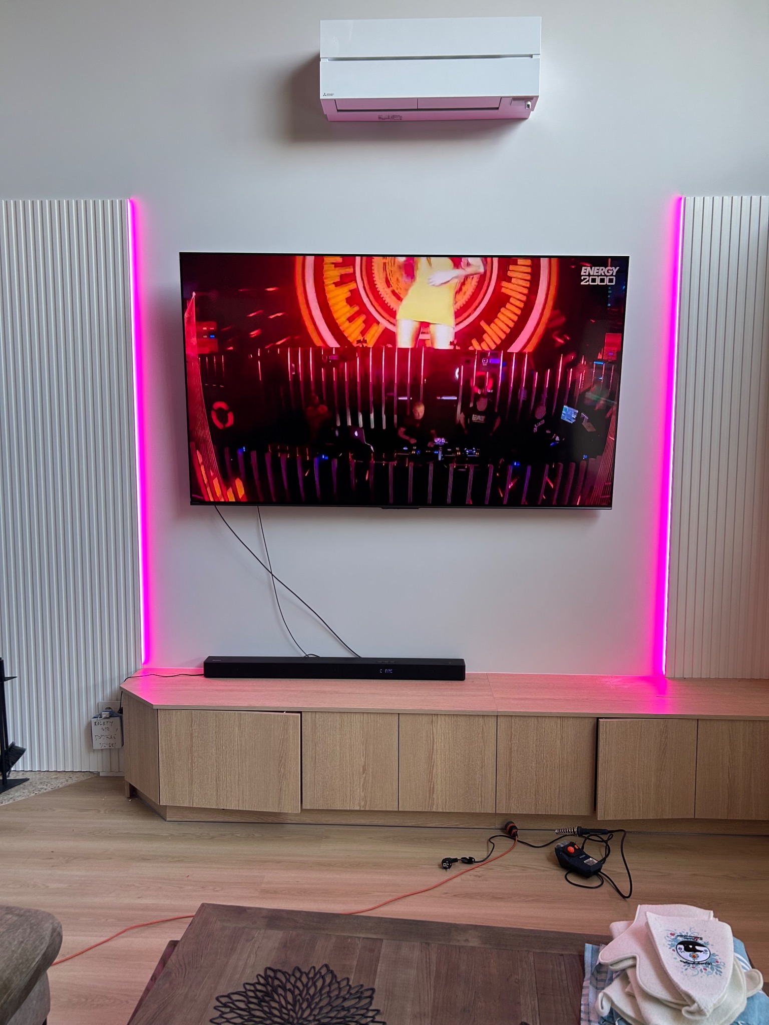 Nowoczesny salon z podświetlanymi panelami ściennymi po bokach telewizora. Na ścianie klimatyzator. Na szafce RTV leży soundbar. Widoczne kable i akcesoria.