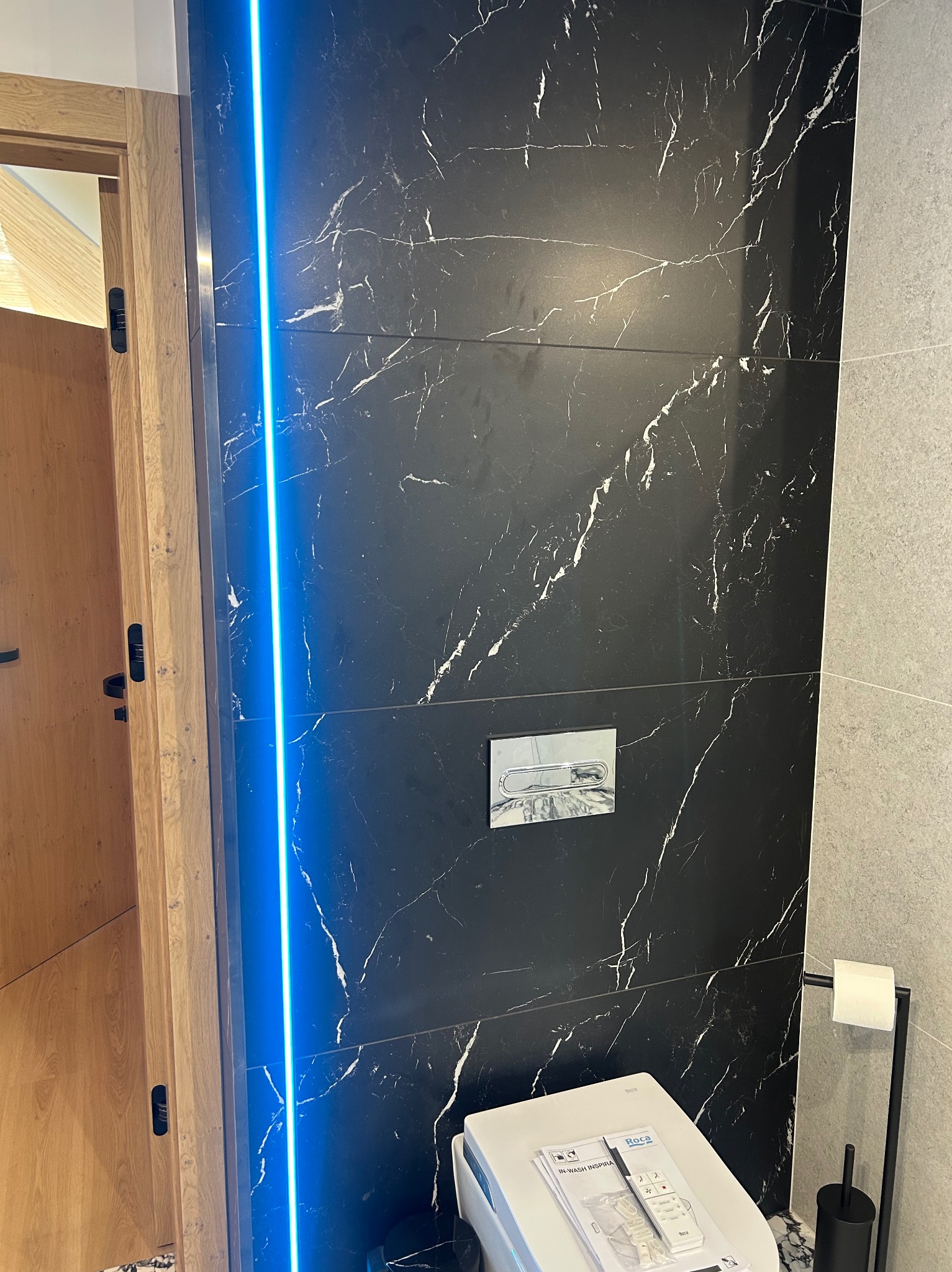 Nowoczesna toaleta z podświetleniem LED i czarnymi płytkami imitującymi marmur. Widoczny drewniany futryna i uchwyt na papier toaletowy. Minimalistyczny design.