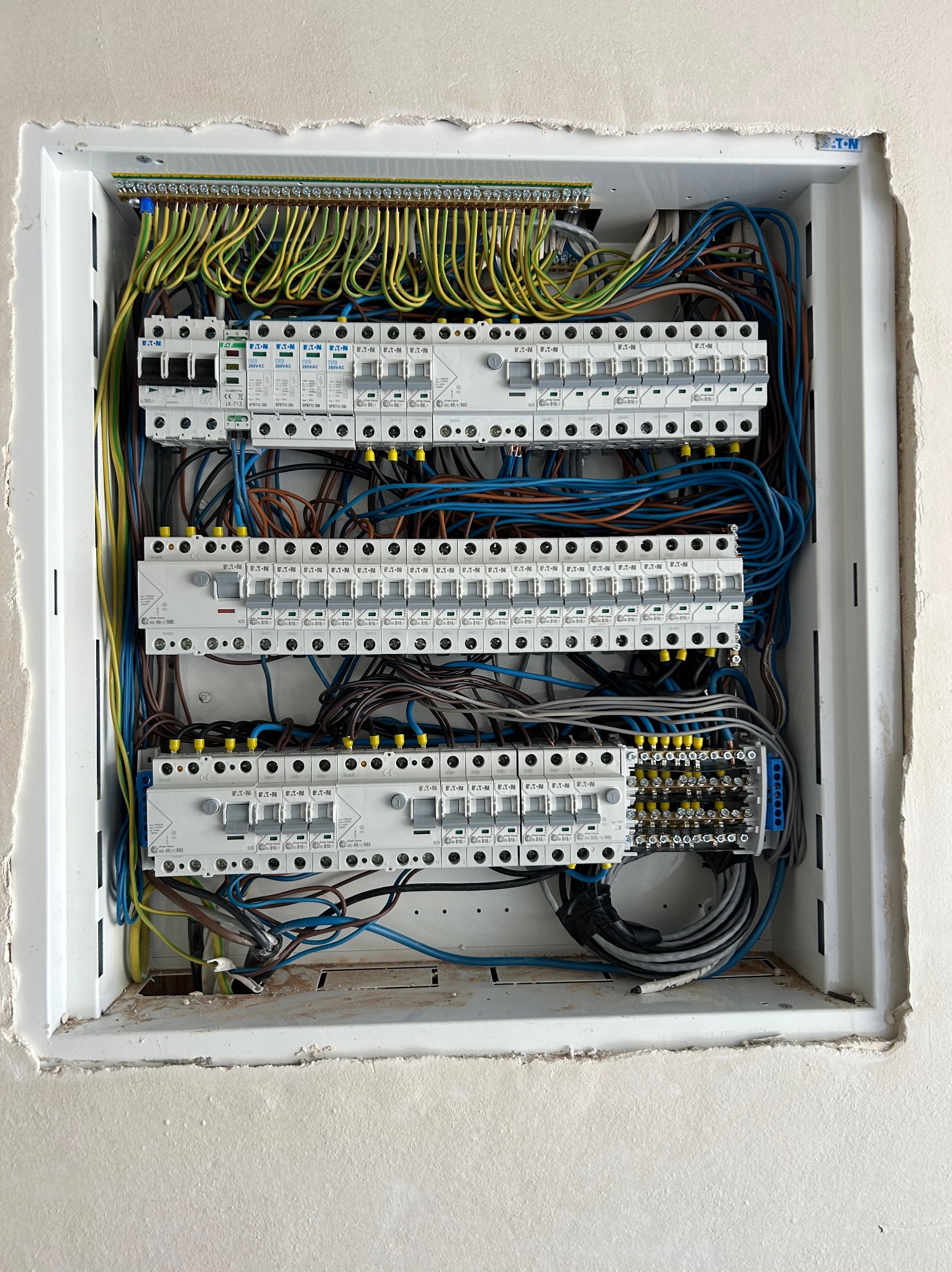 Rozdzielnica elektryczna po montażu, widoczne zabezpieczenia EATON, kable w kolorach: żółto-zielonym, niebieskim, brązowym i czarnym. Biała obudowa w ścianie.