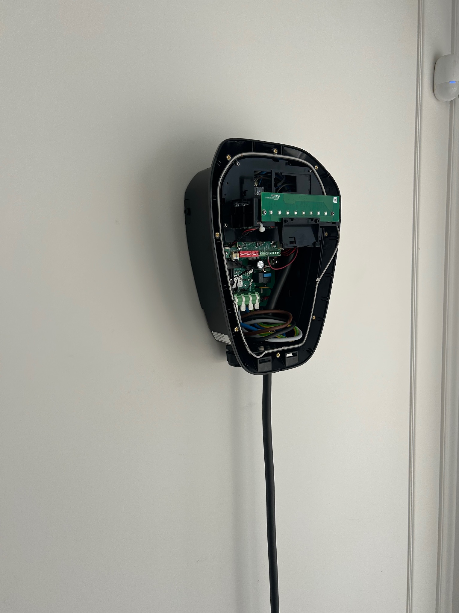 Rozmontowana, czarna stacja ładowania EV na białej ścianie, z widocznymi wewnętrznymi obwodami i okablowaniem. Biały czujnik alarmowy w tle, obok framugi drzwi.