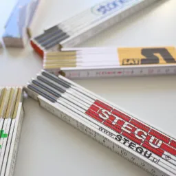 Rozłożone, składane drewniane miarki z nadrukowanymi logo firm budowlanych, ułożone w okrąg na białym tle, uzupełnione o ołówki.