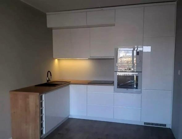 Biała, nowoczesna zabudowa kuchenna z drewnianym blatem, oświetleniem podszafkowym i wbudowanym piekarnikiem, widok na miasto przez szybę piekarnika.