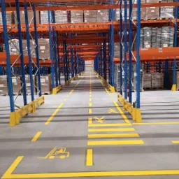 Pełny magazyn z wysokimi regałami w kolorach pomarańczowym i niebieskim, wypełnionymi zapakowanymi towarami, z żółtymi liniami i symbolami na podłodze wyznaczającymi ścieżki transportowe...