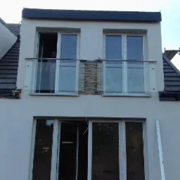 Dwupiętrowy budynek w trakcie wykończenia z balkonem z szklaną balustradą i fragmentem dekoracyjnej ściany z kamienia, widoczny pracownik w oknie i rusztowanie z boku.