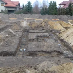 Zbrojenie fundamentów w wykopie, widoczne drewniane znaczniki, w tle dwa domy jednorodzinne w stanie surowym i hałdy ziemi.