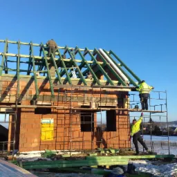 Wznoszenie szkieletu dachu na surowym budynku z cegły ceramicznej, pracownicy na rusztowaniu, w tle ośnieżony krajobraz i błękitne niebo.