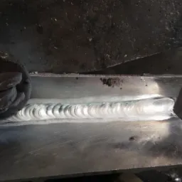 Spoina na metalowej konstrukcji, widoczna krawędź spawu z charakterystycznym wzorem, uchwyt zaciskowy po lewej stronie, surowe otoczenie warsztatowe.