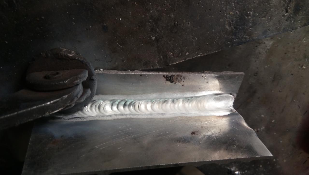 Spoina na metalowej konstrukcji, widoczna krawędź spawu z charakterystycznym wzorem, uchwyt zaciskowy po lewej stronie, surowe otoczenie warsztatowe.