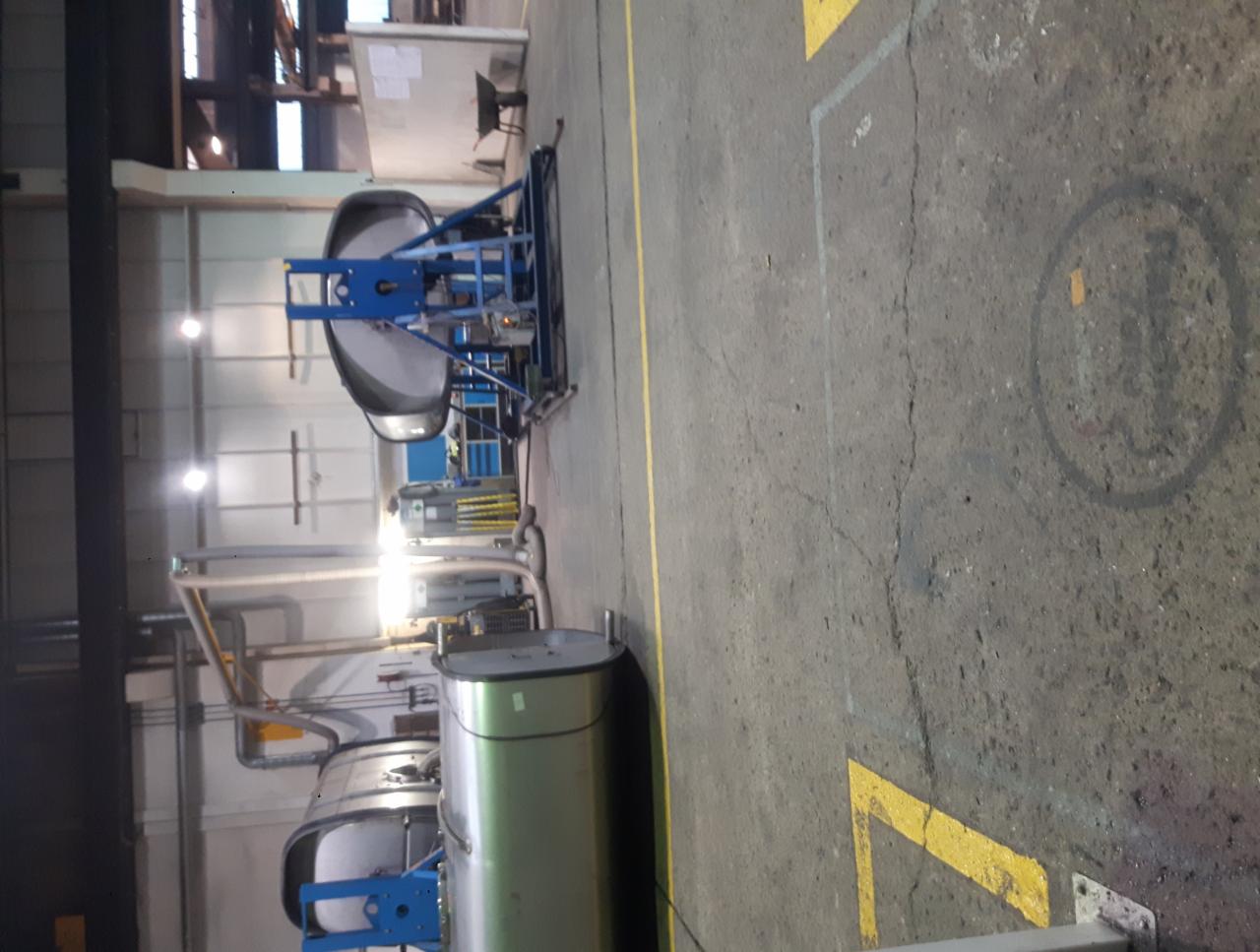 Wnętrze hali produkcyjnej z dwoma metalowymi zbiornikami na niebieskich wózkach, widoczne rury i instalacje pod sufitem, betonowa posadzka z żółtymi oznaczeniami.