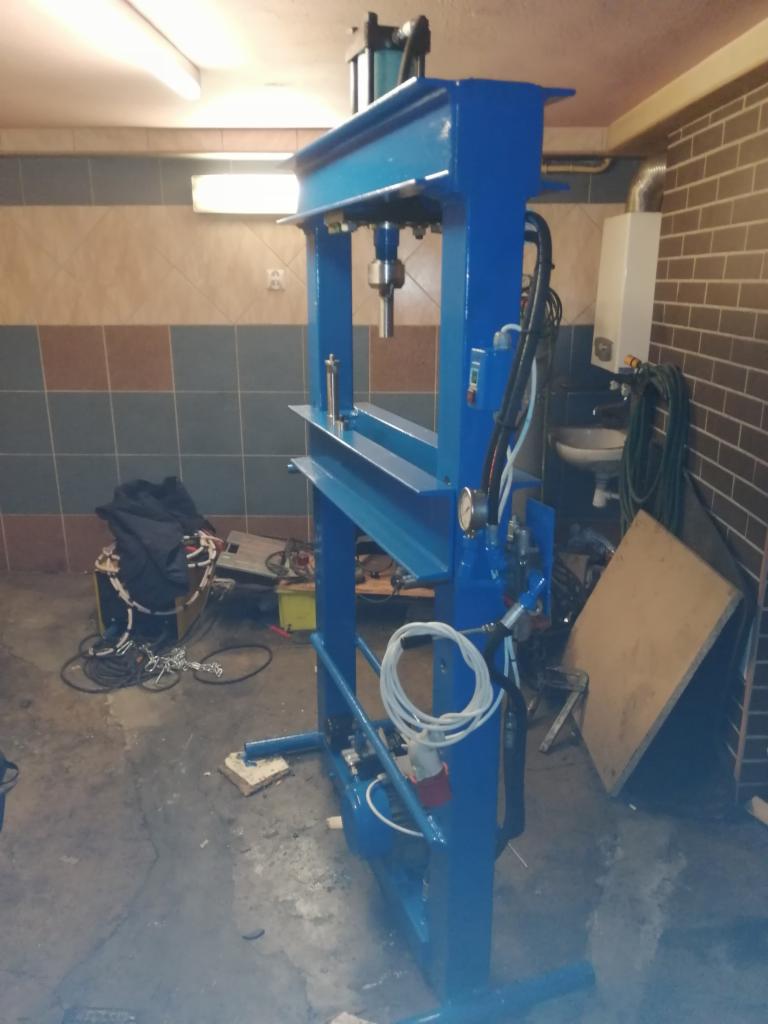 Niebieska prasa hydrauliczna w warsztacie z widocznymi przewodami, manometrem i siłownikiem pneumatycznym.