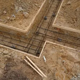 Zbrojenie fundamentów w wykopie z drewnianym szalunkiem, widok z góry na ziemię i stalowe pręty połączone w siatkę.