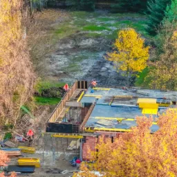 Budowa domu murowanego w Warszawie, widok z góry w jesiennej scenerii. Ekipa w pomarańczowych kamizelkach pracuje nad fundamentami i szalunkami, otoczona kolorowymi drzewami.