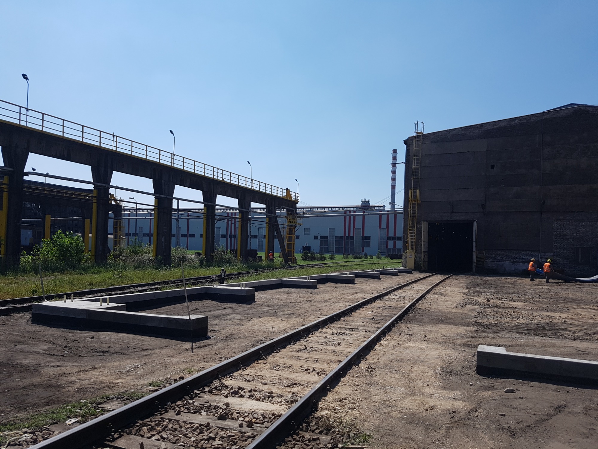 Widok na teren budowy z torami kolejowymi, nowymi betonowymi fundamentami pod konstrukcję i budynkiem przemysłowym w tle, dwóch robotników w pomarańczowych kamizelkach w pobliżu wejścia do budynku.