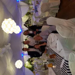 Grupa osób, w tym muzyk z gitarą elektryczną, pozuje do zdjęcia przy udekorowanym stole weselnym pod białym baldachimem z wiszącymi lampami.