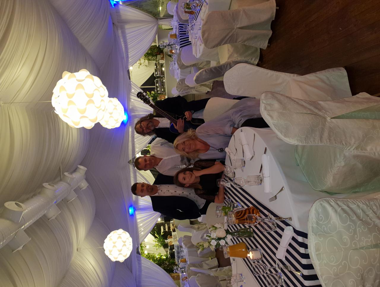 Grupa osób, w tym muzyk z gitarą elektryczną, pozuje do zdjęcia przy udekorowanym stole weselnym pod białym baldachimem z wiszącymi lampami.