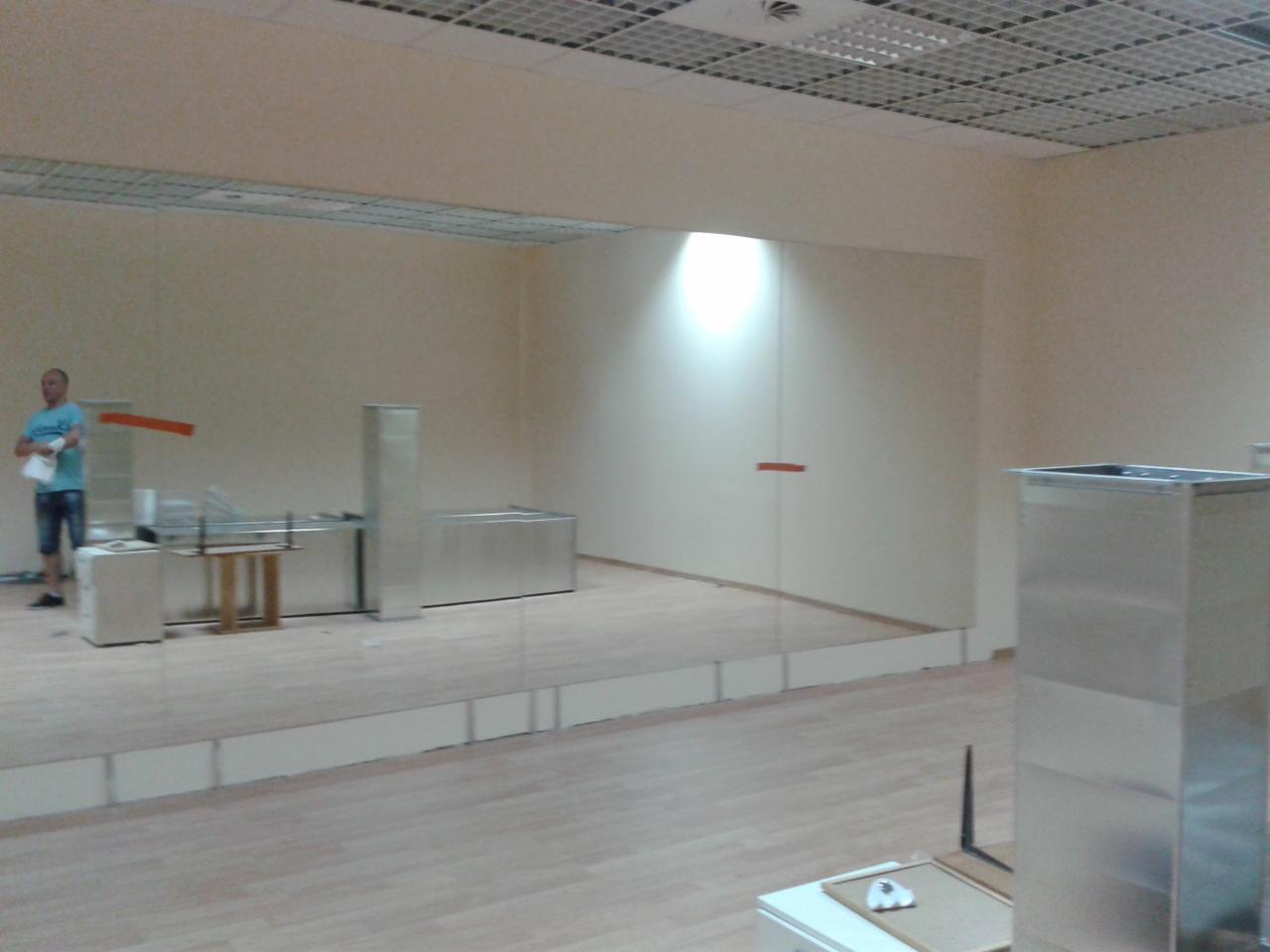 Duże lustro zamontowane na ścianie w jasnym pomieszczeniu z drewnianą podłogą, widoczne elementy instalacji wentylacyjnej i mężczyzna w tle.