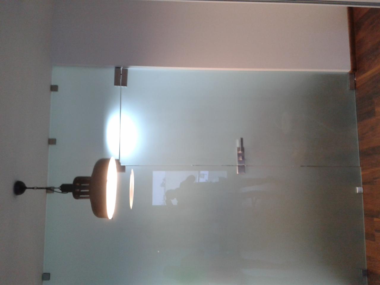 Szklana ściana działowa z matowego szkła, zamocowana na metalowych zawiasach, oświetlona lampą wiszącą, widoczna na tle drewnianej podłogi.