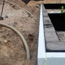 Ocieplenie fundamentu styropianem, widoczna izolacja pionowa oraz rura drenażowa w wykopie.