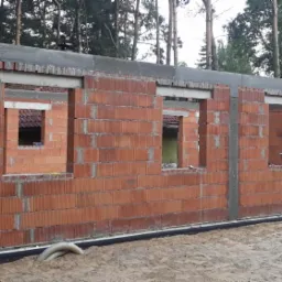 Wznoszenie ścian parterowego budynku z czerwonej ceramiki budowlanej, widoczne otwory okienne i zbrojony wieniec, teren budowy z piaskiem i drzewami w tle.