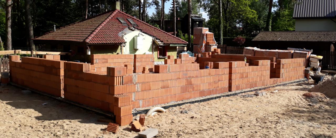Wznoszone ściany z czerwonej cegły na fundamencie domu jednorodzinnego w trakcie budowy, widoczne pustaki i przygotowane materiały budowlane, w tle zalesiony teren.
