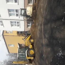 Żółta koparko-ładowarka Komatsu oraz minikoparka JCB na placu budowy, w tle budynek mieszkalny z białymi oknami, widoczne świeże ślady po wykopie.